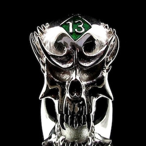 Może przedstawiać: Srebrny pierścionek z czaszką, na którym znajduje się zielony kamień szlachetny. Kamień ma na sobie cyfrę 13.