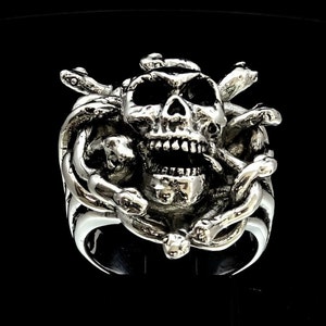 May include: A silver ring with a detailed skull design surrounded by snakes. The skull has an open mouth and a tongue sticking out.