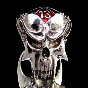 May include: A silver skull ring with a red enamel diamond containing the number "13". The skull has sharp teeth and horns.