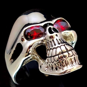 Op de afbeelding: Een zilveren schedelring met rode edelsteenogen. De schedel is gedetailleerd met een getextureerd oppervlak en een tandenrij.