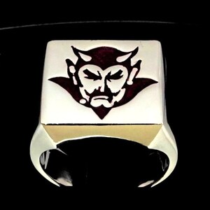 May include: A silver ring with a square face featuring a red devil head graphic.