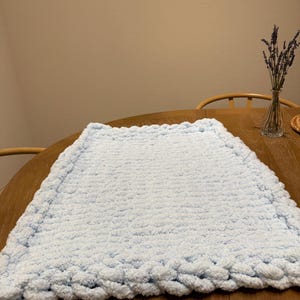 May include: A light blue, hand-knitted blanket with a braided edge. The blanket is made of a soft, plush material and is displayed on a wooden table. A vase of lavender is in the background.