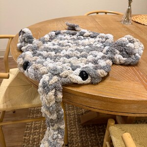 May include: A handmade, plush crocheted stingray in shades of gray, beige, and white. The toy features large, black button eyes and a long, dangling tail. The stingray is resting on a wooden table.