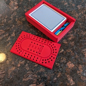 Peut inclure: Un plateau de jeu en plastique rouge avec une zone de départ circulaire et des cases numérotées. Le plateau est conçu pour un jeu de cribbage. Le plateau est ouvert et une boîte en plastique rouge avec une carte à l'intérieur est à côté.