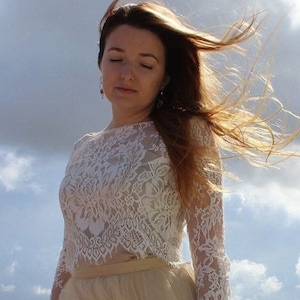 May include: A white lace top with long sleeves and a round neckline. The intricate floral lace design covers the entire top. The model is wearing a beige skirt. The background is a cloudy sky.