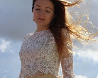 Op de afbeelding: Een witte kanten top met lange mouwen en een ronde halslijn. Het ingewikkelde bloemenkantontwerp bedekt de hele top. Het model draagt een beige rok. De achtergrond is een bewolkte lucht.