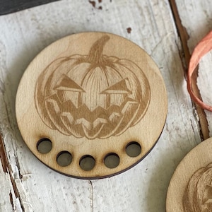 May include: A wooden pumpkin-shaped yarn bowl with five holes for holding yarn. The pumpkin has a carved face with a mischievous grin.