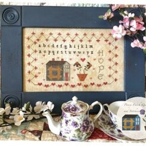 May include: Cross stitch pattern featuring a house with a heart and a flower pot with two flowers. The text "Hope" is stitched below the flower pot. The alphabet is stitched above the house. The pattern is framed in a blue frame.