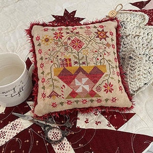May include: A square, beige decorative pillow with a cross-stitch floral design in red, yellow, and green. The pillow has a red fringe and is next to a white mug with the text "merry & bright". A pair of scissors is also visible.