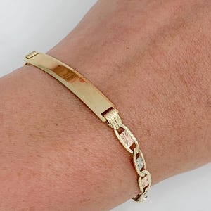 May include: A gold-colored bracelet with a rectangular nameplate and a chain-link design. The bracelet is worn on a wrist, showcasing its polished finish and intricate details. The jewelry piece is a classic accessory.