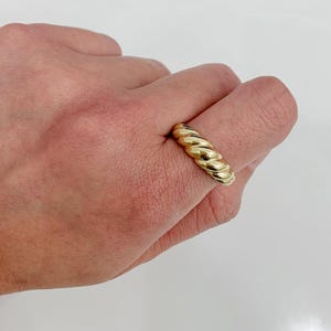 May include: A gold-colored ring with a twisted rope design. The ring is shown on a hand against a white background. The ring's design features a series of rounded ridges, creating a textured appearance. The ring is a simple yet elegant piece of jewelry.
