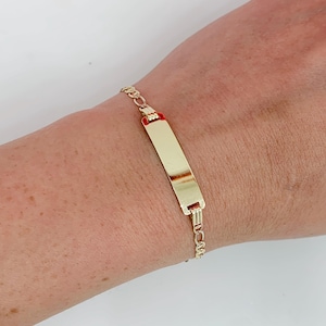 May include: A gold ID bracelet featuring a rectangular plate and a delicate chain. The plate has a polished finish, and the chain has small, interconnected links. The bracelet is worn on a wrist.