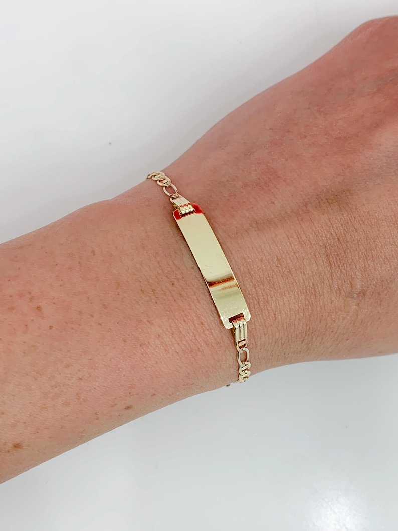 May include: A gold ID bracelet featuring a rectangular plate and a delicate chain. The plate has a polished finish, and the chain has small, interconnected links. The bracelet is worn on a wrist.