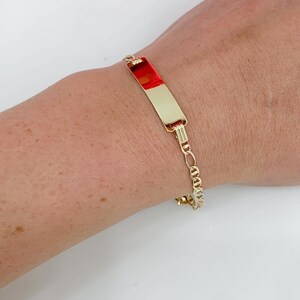 May include: Gold-coloured bracelet with a rectangular plate featuring a red and gold design. The bracelet has a chain link design and is worn on a wrist.