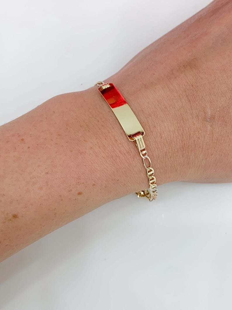 May include: Gold-coloured bracelet with a rectangular plate featuring a red and gold design. The bracelet has a chain link design and is worn on a wrist.