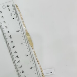 May include: A gold bracelet featuring a rectangular nameplate and a delicate chain. The bracelet is measured with a ruler, showing a length of approximately 15 cm. The bracelet is set against a white background.