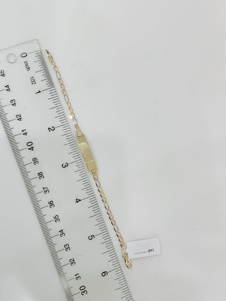 May include: A gold bracelet featuring a rectangular nameplate and a delicate chain. The bracelet is measured with a ruler, showing a length of approximately 15 cm. The bracelet is set against a white background.