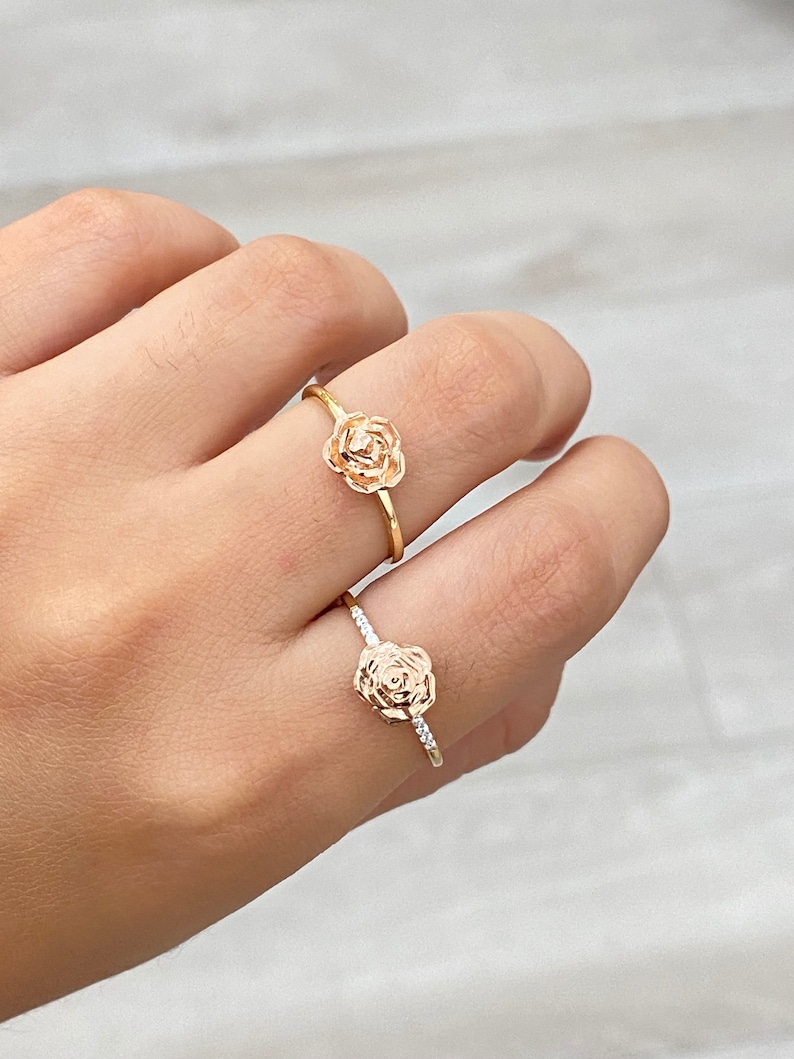 May include: Two gold rose rings. The ring on the top has a plain band. The ring on the bottom has a band with small clear stones.