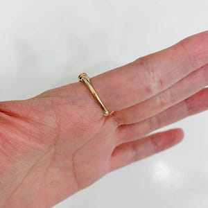 May include: A gold ring with a small, round, colorful stone. The ring is set on a finger, showcasing its simple yet elegant design. The band is thin and polished, reflecting light.
