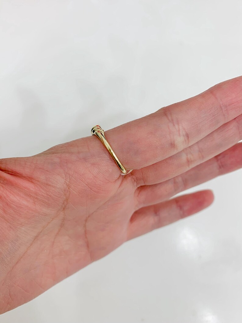 May include: A gold ring with a small, round, colorful stone. The ring is set on a finger, showcasing its simple yet elegant design. The band is thin and polished, reflecting light.