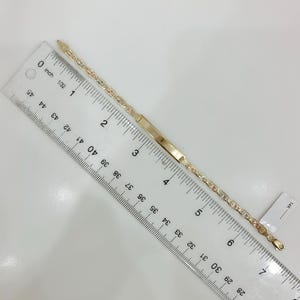 May include: A gold bracelet with a rectangular nameplate and a delicate chain design. The bracelet is approximately 7 inches (17.78 cm) long and features a mix of gold colors. A ruler is placed next to the bracelet.
