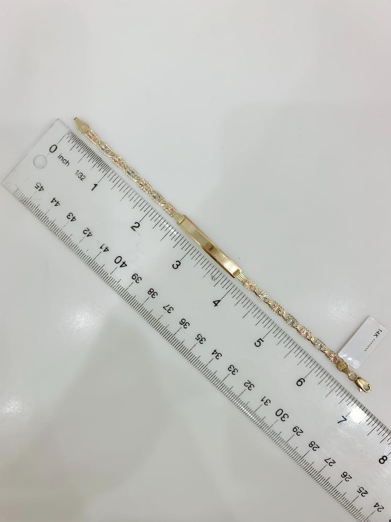 May include: A gold bracelet with a rectangular nameplate and a delicate chain design. The bracelet is approximately 7 inches (17.78 cm) long and features a mix of gold colors. A ruler is placed next to the bracelet.