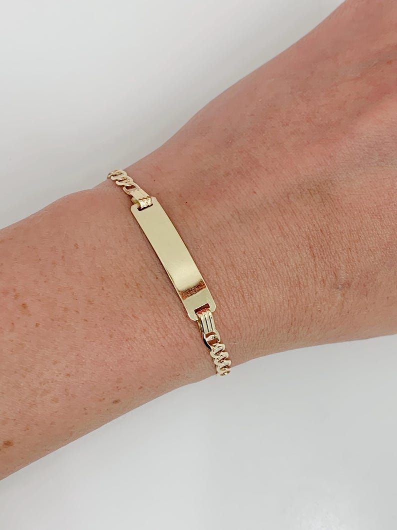 May include: A gold-colored bracelet with a rectangular nameplate and a curb chain design. The bracelet is shown on a wrist, and the metal has a polished finish. The nameplate is blank, ready for engraving.