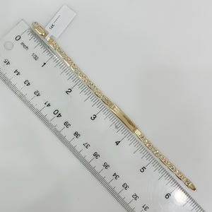 May include: A gold-colored bracelet with a rectangular bar and a line of small, clear stones. The bracelet is measured with a ruler, showing a length of approximately 6 inches. A tag with "14K" is attached.