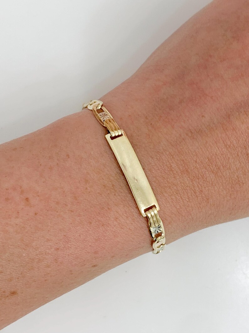 May include: Gold-tone bracelet with a rectangular center plate and chain links. The center plate is plain, while the chain links feature star-like designs. The bracelet is worn on a wrist.