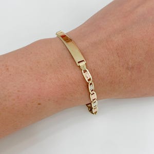 May include: A gold-toned ID bracelet with a rectangular plate and a chain-link design. The bracelet is worn on a wrist, showcasing its polished finish and detailed links. The bracelet is a classic piece of jewelry.