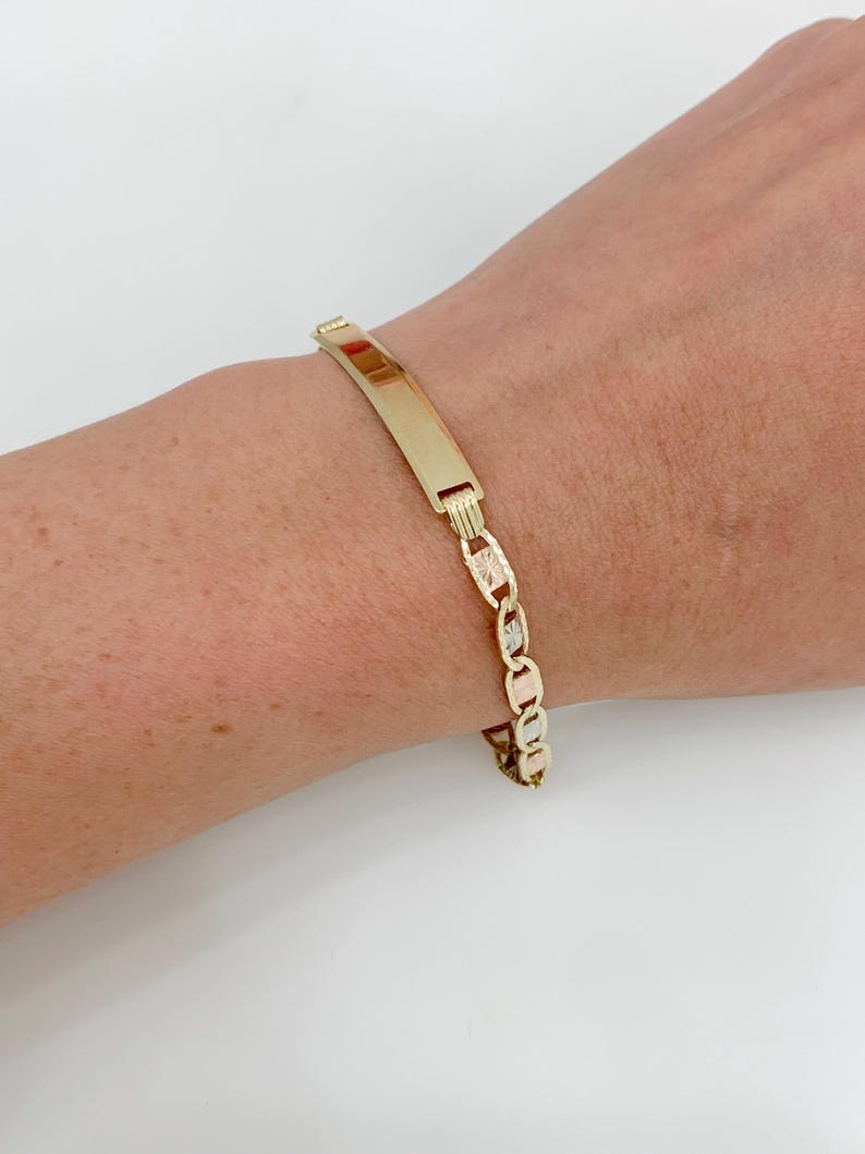 May include: A gold-toned ID bracelet with a rectangular plate and a chain-link design. The bracelet is worn on a wrist, showcasing its polished finish and detailed links. The bracelet is a classic piece of jewelry.