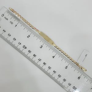 May include: A gold-colored ID bracelet with a rectangular plate and a chain link design. The bracelet is measured with a ruler, showing a length of approximately 6 inches. A small tag with "14K" is visible.