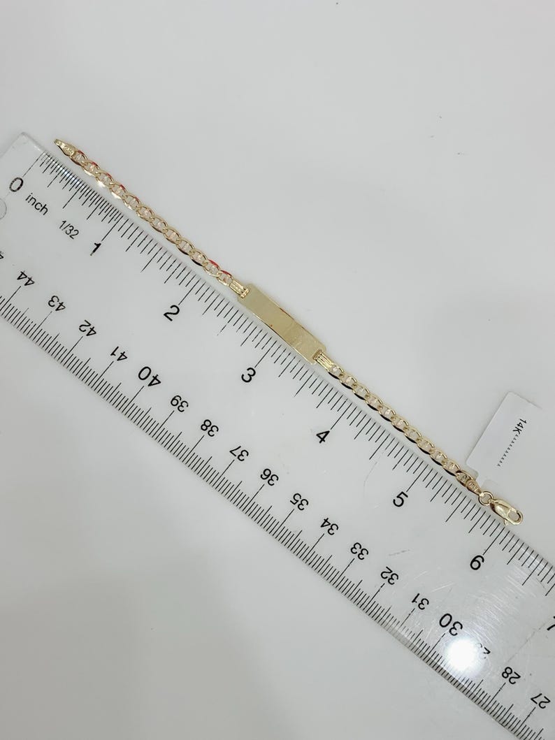 May include: A gold-colored ID bracelet with a rectangular plate and a chain link design. The bracelet is measured with a ruler, showing a length of approximately 6 inches. A small tag with "14K" is visible.
