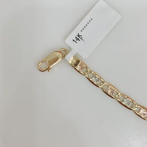 May include: A gold-colored chain bracelet with a lobster clasp. The chain links feature a starburst design with alternating gold, silver, and rose gold tones. A white tag with "14K" is attached.