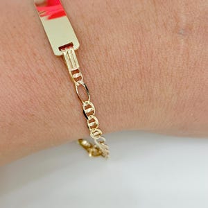 May include: A gold ID bracelet. The rectangular ID plate has a polished finish with a red and pink reflection. The bracelet features a Figaro chain design. The bracelet is on a wrist.