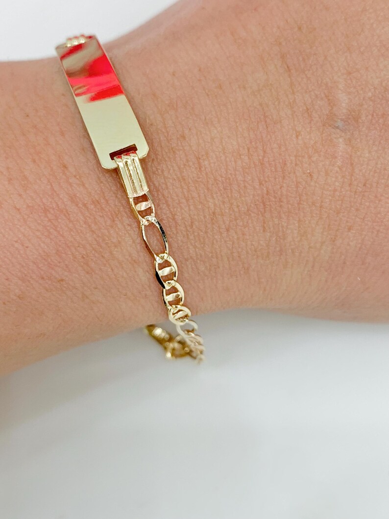 May include: A gold ID bracelet. The rectangular ID plate has a polished finish with a red and pink reflection. The bracelet features a Figaro chain design. The bracelet is on a wrist.