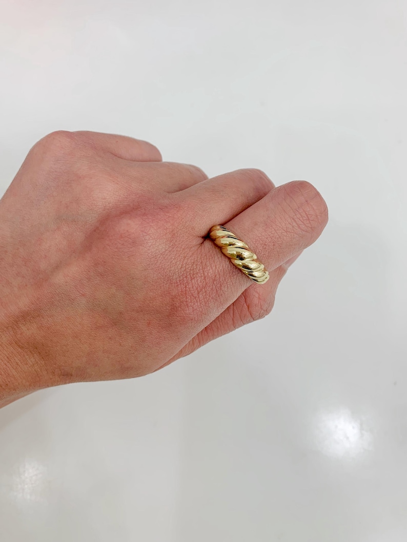 May include: A gold-colored ring with a twisted rope design. The ring is shown on a hand against a white background. The ring's design features a series of rounded ridges, creating a textured appearance. The ring is a simple yet elegant piece of jewelry.