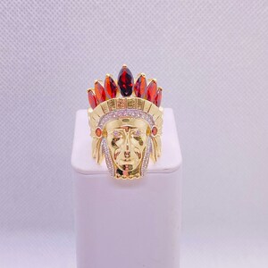 May include: Gold ring with a detailed Native American chief head design. The ring is adorned with red and clear gemstones.