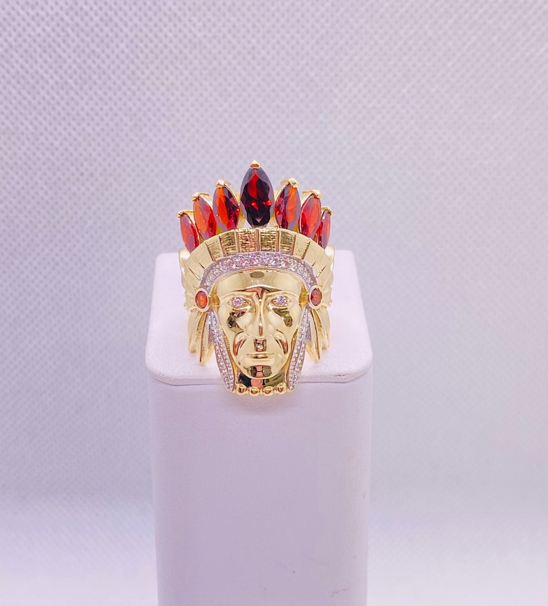 May include: Gold ring with a detailed Native American chief head design. The ring is adorned with red and clear gemstones.