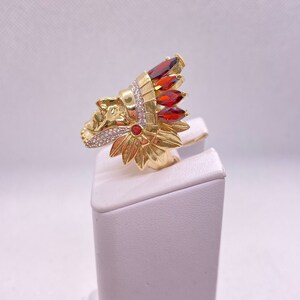 May include: Gold ring with a detailed design of an Native American chief's head wearing a headdress with red and white stones.