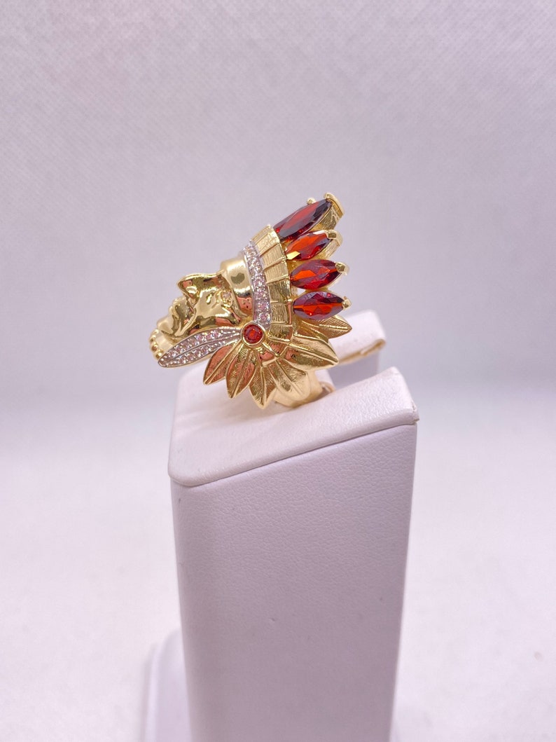 May include: Gold ring with a detailed design of an Native American chief's head wearing a headdress with red and white stones.