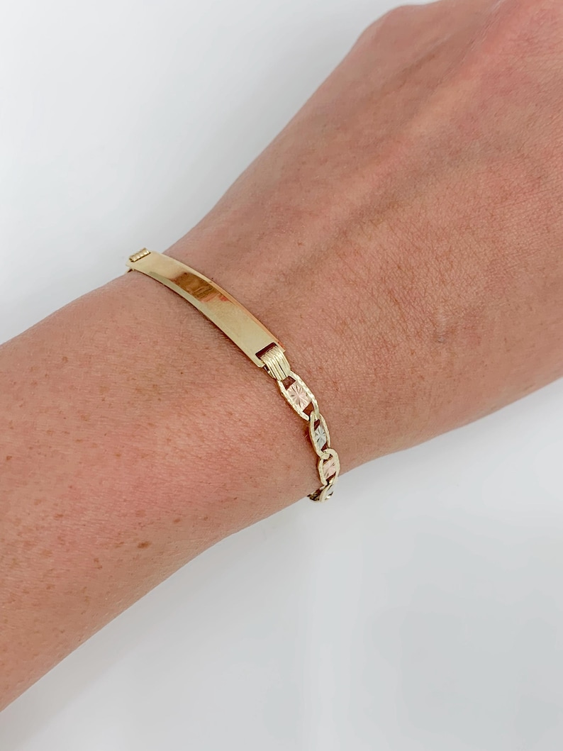 May include: A gold-colored bracelet with a rectangular nameplate and a chain-link design. The bracelet is worn on a wrist, showcasing its polished finish and intricate details. The jewelry piece is a classic accessory.