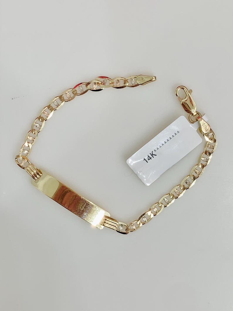 May include: A gold-colored bracelet with a rectangular nameplate and a chain link design. The bracelet has a lobster clasp closure and a small tag that reads "14K". The bracelet is displayed on a white surface.