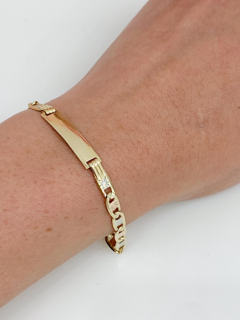 May include: A gold-colored bracelet with a rectangular nameplate and a chain-link design. The bracelet features a small, square-shaped embellishment with a clear stone. The bracelet is worn on a wrist.