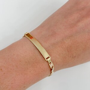 May include: A gold-colored ID bracelet with a rectangular plate and a chain link band. The bracelet is worn on a wrist against a white background. The plate is smooth and shiny.