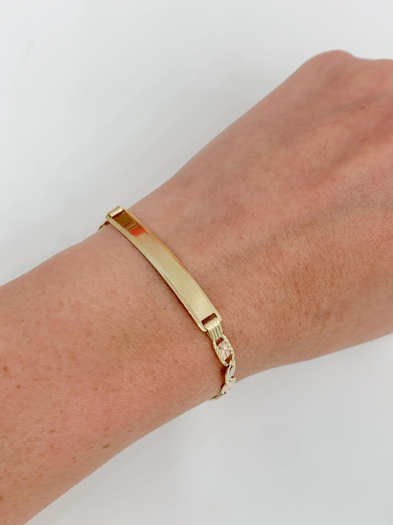 May include: A gold-colored ID bracelet with a rectangular plate and a chain link band. The bracelet is worn on a wrist against a white background. The plate is smooth and shiny.