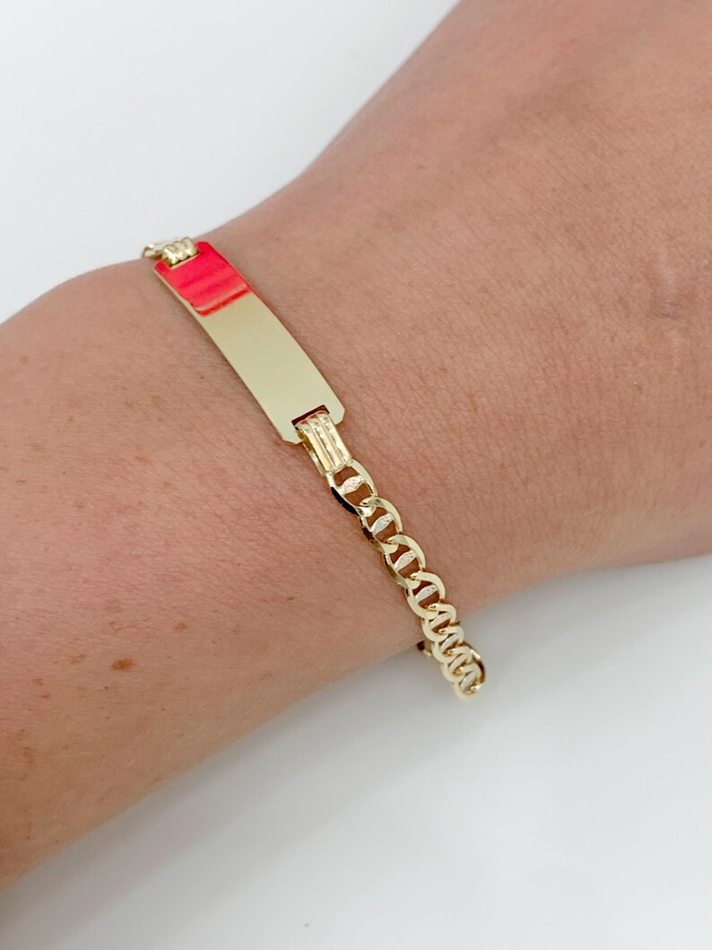 May include: A gold-colored bracelet with a rectangular nameplate and a chain link design. The nameplate is smooth and polished, and the chain links are small and connected. The bracelet is worn on a wrist.