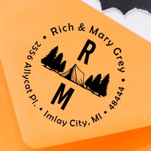 May include: An orange envelope with a black circular stamp. The stamp features a tent, trees, and the initials "RM". Text around the circle reads "Rich & Mary Grey", "2556 Allycat Pl.", and "Imlay City, MI 48444".