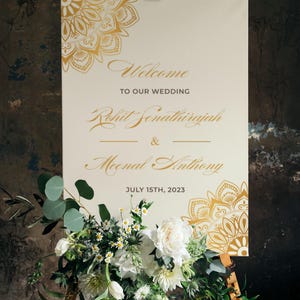 Op de afbeelding: Een welkomstbord voor een bruiloft op een ezel. Het crèmekleurige bord heeft gouden bloemmotieven en de tekst "Welkom op onze bruiloft", gevolgd door namen en de datum 15 juli 2023. Een bloemstuk van witte bloemen en groen siert de basis.