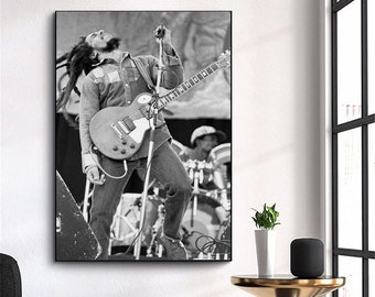 May include: Black and white photo of a musician playing a guitar on stage. The musician is wearing a long-sleeved shirt and jeans. The guitar is a solid body electric guitar with a single cutaway.